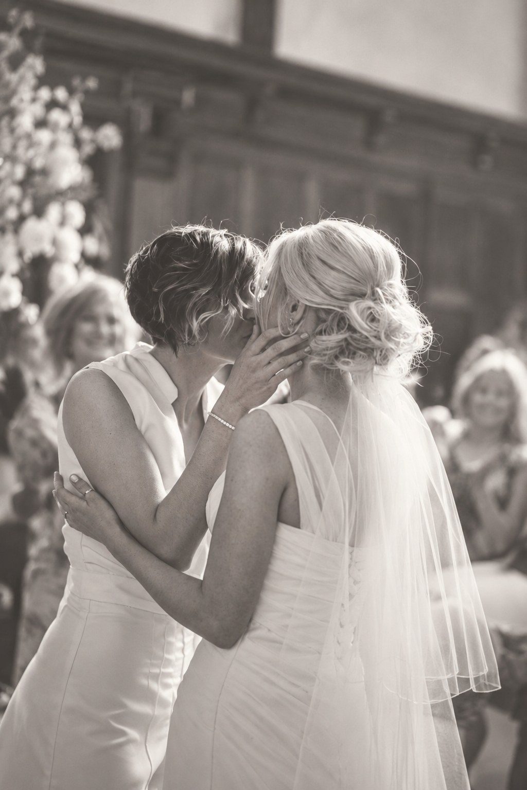 Two brides sharing a kiss at their ceremony as smiling guests look on, in soft sepia