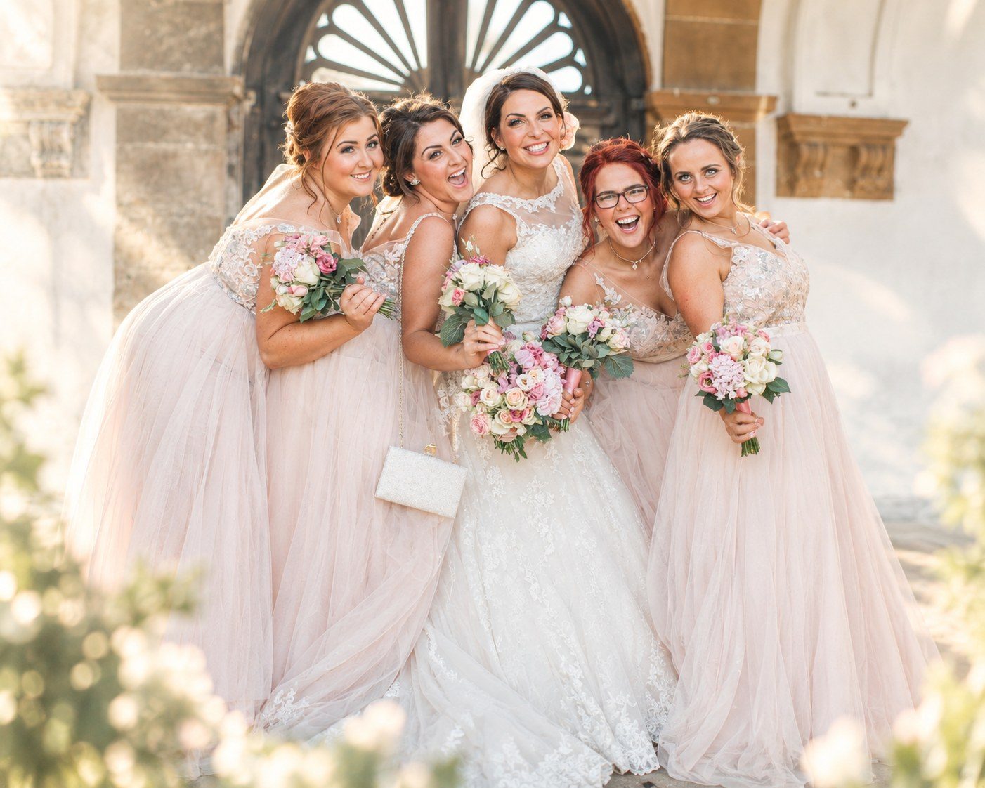 Bride laughing with four bridesmaids in blush tulle dresses holding pink bouquets in warm sunlight