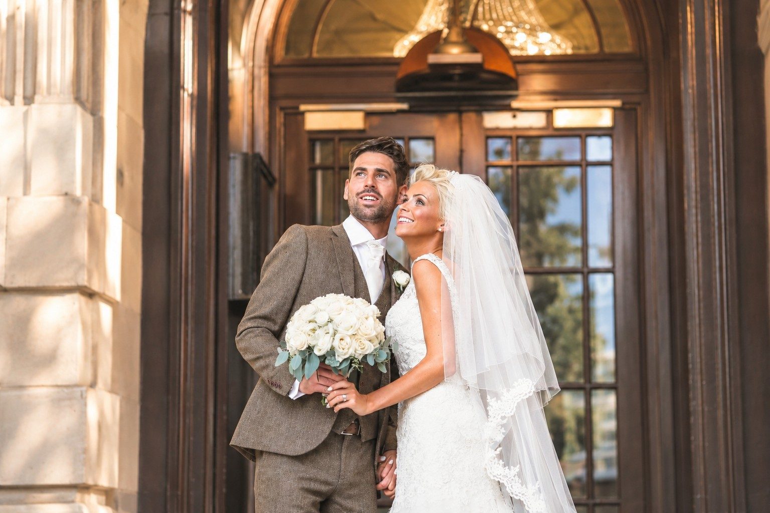 Bride and groom smiling up together in a grand wooden doorway, holding a bouquet of white roses