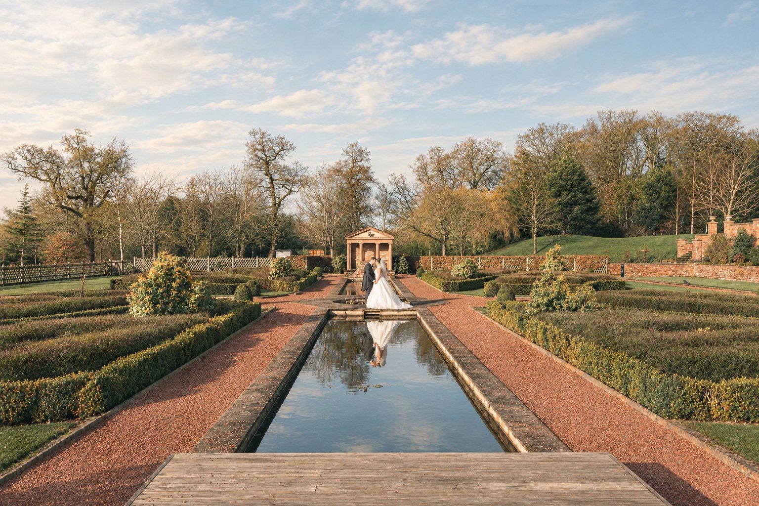 Bride and groom kissing at the end of a long reflecting pool in a formal garden, their reflection in the water