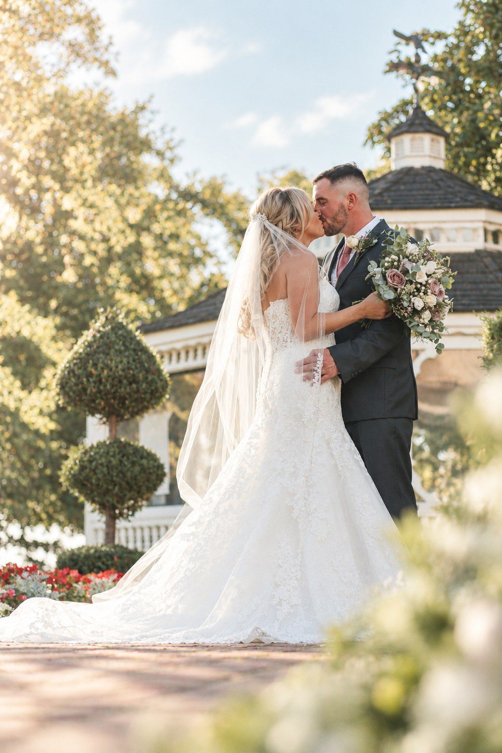 Bride and groom kissing in front of a garden gazebo in warm sunlight, her long veil and lace train flowing