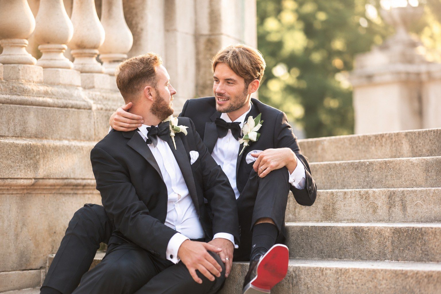 Two grooms in black tie sitting close on stone steps, smiling at each other