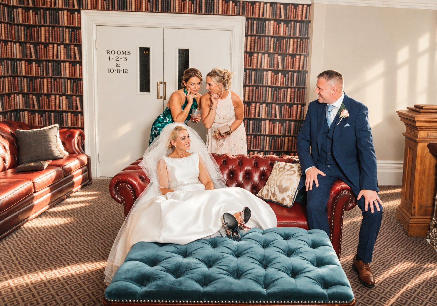 Bride relaxing on a red leather sofa with her feet up, friends chatting behind her and the groom smiling beside her in a library room