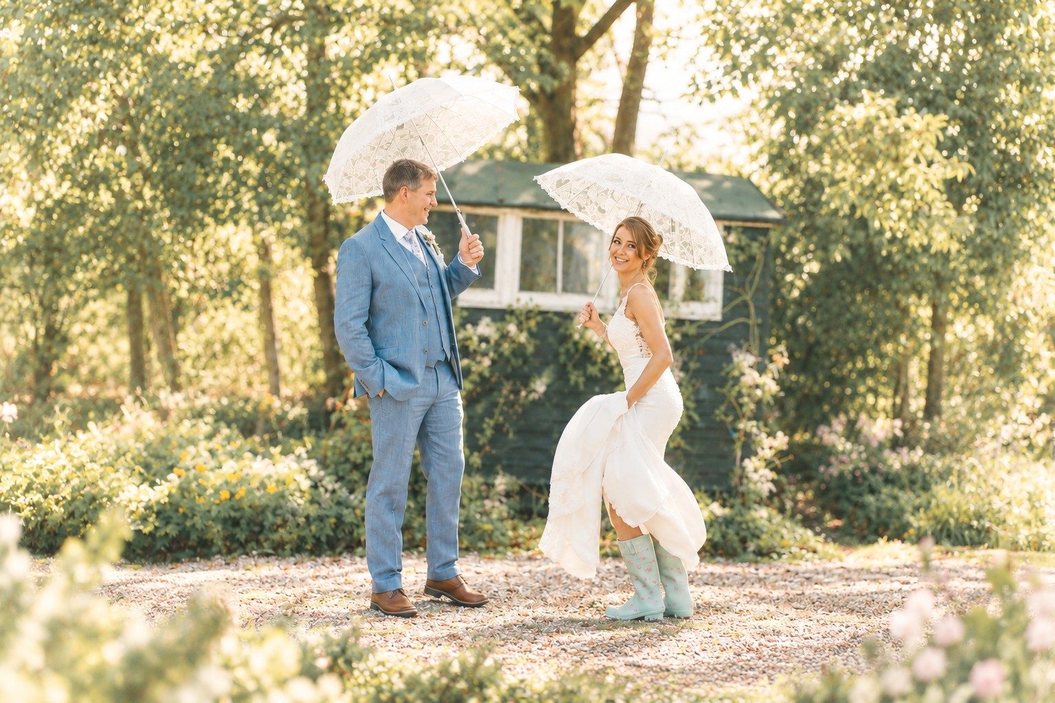 Bride in floral wellies and groom in a blue suit laughing under white lace umbrellas in a sunlit garden
